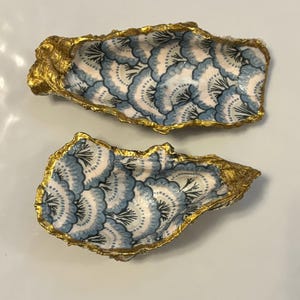 以下が含まれることがあります： 金色の縁取りが施された2つの装飾的なカキ殻。各殻の内側には、青と白の花柄が施されています。これらの殻は白い表面に配置されており、そのユニークなデザインと職人技が際立っています。
