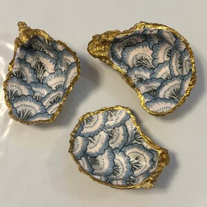 以下が含まれることがあります： 金色の縁取りが施された、3つの装飾的なカキの貝殻。貝殻には青と白の花柄が描かれています。貝殻は白い表面に配置されています。