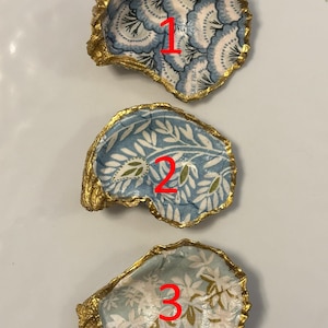 以下が含まれることがあります： 金色の縁取りが施された、3つの装飾的なカキ殻。それぞれ、青、白、金色のユニークな模様が内側に施されています。白い表面に配置されています。