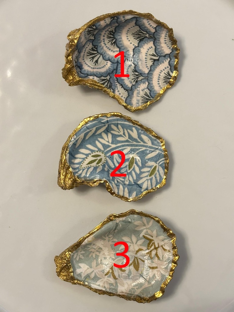 以下が含まれることがあります： 金色の縁取りが施された、3つの装飾的なカキ殻。それぞれ、青、白、金色のユニークな模様が内側に施されています。白い表面に配置されています。