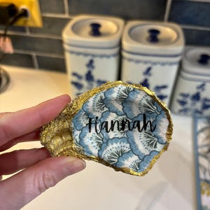 May include: A handcrafted oyster shell with a gold-painted edge. The interior features a blue and white floral pattern and the name "Hannah" in black script. A unique decorative item.