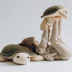 Pode incluir: Uma criança usando um chapéu de tricô marrom e um suéter bege brinca com dois brinquedos de pelúcia de tartaruga. Uma tartaruga é verde e a outra é bege com uma casca verde.