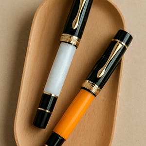 May include: Two fountain pens with black and gold accents rest on a wooden tray. One pen has a white barrel, the other an orange barrel. Both pens feature gold-colored clips and decorative bands, creating a classic and elegant aesthetic.