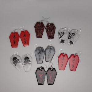 Könnte beinhalten: Eine Sammlung von Sarg-Ohrringen zum Thema Halloween in verschiedenen Farben und Designs. Einige Ohrringe sind rot, grau oder schwarz, mit Glitzer oder grafischen Elementen wie Totenköpfen, Spinnen und den Buchstaben "RIP".