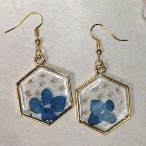 Peut inclure: Une paire de boucles d'oreilles hexagonales dorées. Chaque boucle d'oreille présente une fleur bleue enfermée dans de la résine transparente, avec de petites fleurs blanches et des paillettes. Les boucles d'oreilles ont des crochets d'oreille dorés.
