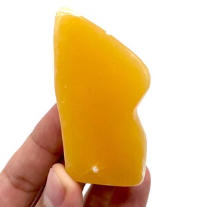 May include: A smooth, orange-yellow, handheld stone with a curved shape and a small indentation. The stone is held in a person's hand against a white background. The stone appears to be a gua sha tool.