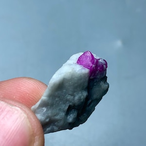 May include: A close-up of a small, rough-hewn rock with a cluster of vibrant, hot pink crystals. The crystals are sharply defined against the light gray stone, creating a striking contrast. The rock is held by a person's fingers.
