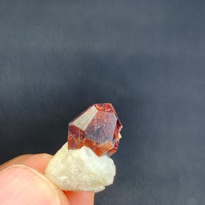 Puede incluir: Un cristal mineral facetado de color marrón rojizo se asienta sobre una roca blanca y porosa. El cristal tiene una forma geométrica con bordes afilados y una superficie brillante. El fondo es de color gris oscuro.