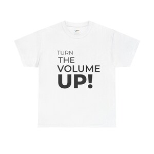 Puede incluir: Camiseta blanca con la frase "TURN THE VOLUME UP!" impresa en negro. El texto está centrado en la parte delantera de la camiseta. La camiseta tiene mangas cortas y cuello redondo.