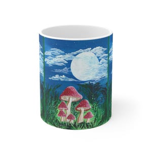 Op de afbeelding: Witte keramische mok met een bosscène-ontwerp. Het kunstwerk toont rood-wit gevlekte paddenstoelen, hoog groen gras, een volle maan en een sterrenhemel. De mok heeft een glanzende afwerking en een standaard handvat.