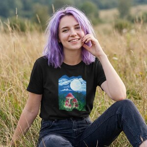 Könnte beinhalten: Schwarzes T-Shirt mit einer farbenfrohen Illustration von roten Pilzen unter einem Vollmond. Das Design ist vor einem Hintergrund aus blauem Himmel und grünem Gras platziert. Die Person trägt eine dunkelblaue Jeans und sitzt in einem Feld mit hohem Gras.
