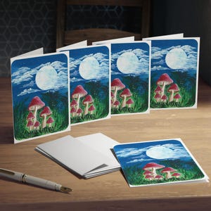 Könnte beinhalten: Eine Reihe von Grußkarten mit einer skurrilen Illustration von rotkappigen Pilzen unter einem Vollmond in einem Nachthimmel. Die Karten sind weiß mit glänzendem Finish, und das Kunstwerk ist lebendig mit Blau-, Grün- und Rottönen.