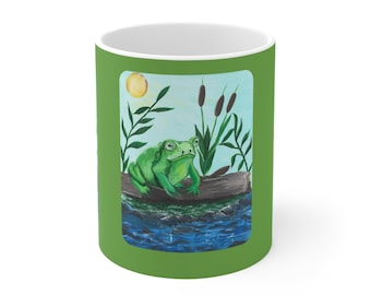 May include: A white ceramic mug with a green exterior featuring a painting of a green frog on a log in a pond. The artwork includes cattails, a yellow sun, and blue water.