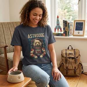 Può includere: T-shirt grigio scuro con un barboncino dei cartoni animati in tuta spaziale e la scritta "ASTRODOG". La grafica è rossa, bianca e blu. La persona indossa jeans blu. Una ciotola per cani con un'impronta di zampa è in primo piano.
