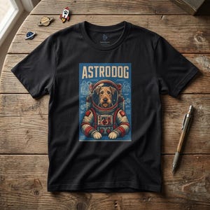 Può includere: T-shirt nera con grafica di un cane in tuta spaziale e la scritta "ASTRODOG". Il cane è marrone e bianco, su sfondo blu. La maglietta è appoggiata su una superficie di legno.