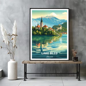 Può includere: Una stampa artistica incorniciata del lago di Bled, in Slovenia, con una chiesa su un'isola nel lago e montagne sullo sfondo. L'opera d'arte è in stile manifesto di viaggio vintage, con il testo "LAKE BLED Slovenia" in basso.