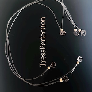 May include: Clear hair extension strands with small metal clips and beige connectors. The image features the text "TressPerfection" in a serif font. The clips are in a variety of shapes and sizes, and the strands are arranged in a circular pattern.