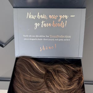 Może przedstawiać: Ciemnoszara skrzynka ze złotymi literami z napisem "New hair, new you - go turn heads!" i "shine!". Wewnątrz pudełka znajduje się brązowa peruka z pasemkami. Tekst brzmi również "Handle with care, Style with love. Your TressPerfection piece is designed to dazzle".