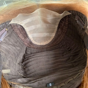 Può includere: Vista interna di una cuffia per parrucca, che mostra la sua costruzione. La cuffia presenta una base in tessuto marrone con un motivo cucito, un frontale in pizzo beige e una toppa in tessuto beige. Le clip metalliche sono visibili lungo i bordi e una piccola "S" in basso.