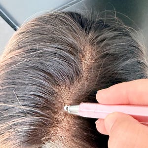 Op de afbeelding: Close-up van een pruik met donkerbruin haar en een kanten voorkant. Een roze tool wordt gebruikt om de pruik aan te passen. De pruik staat op een standaard.