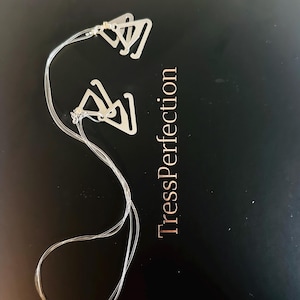 May include: Two silver-colored bra strap clips connected by clear elastic straps. The clips are triangular with a hook design. The text "TressPerfection" is printed in a copper color on a black surface.