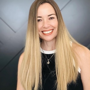 May include: A smiling person with long, blonde hair and a black top with a white trim. A silver necklace with a pendant is visible. The background is a gradient of gray and white.