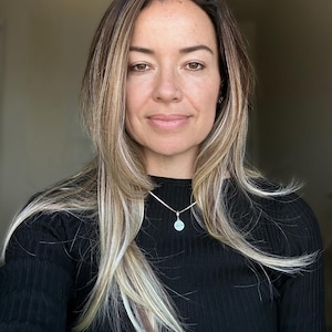 May include: A person with shoulder-length brown hair featuring blonde highlights, wearing a black ribbed long-sleeve shirt and a silver chain necklace with a small, round, light-colored pendant featuring a subtle graphic design.