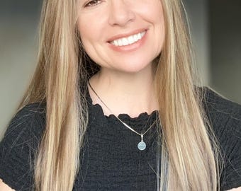 May include: A woman with long blonde hair is wearing a black shirt with a silver necklace and a round pendant. She is smiling at the camera.