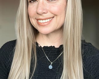 May include: A woman with long, straight, blonde hair is wearing a black, ruffled, short-sleeved shirt. She is wearing a silver necklace with a small, round pendant.