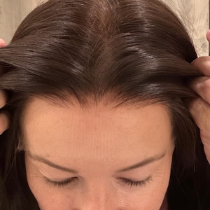 May include: A close-up view of a person's head, showcasing a dark brown wig with a lace front. The wig's hair is styled with a natural part and flows down. The lace front is visible, blending with the person's skin tone.
