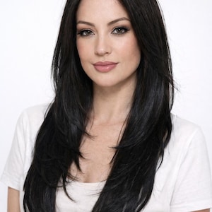 May include: A woman with long, layered black hair and a white t-shirt. The hair is styled with a side part and falls in soft waves. The woman has a fair complexion and is looking at the camera.