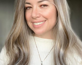 May include: A woman with long, wavy, light brown hair with streaks of gray wears a white sweater and a silver chain necklace with a large, oval-shaped pendant made of agate. The pendant is a dark brown, black, and white agate with a silver bail.