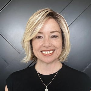 May include: A woman with blonde hair and a black top smiles at the camera. She wears a silver necklace with a small pendant. The background is a dark gray wall with a geometric pattern.
