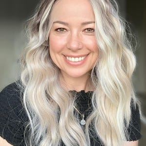 May include: A woman with long, wavy, platinum blonde hair is wearing a black, short-sleeved shirt. She is smiling and has a silver necklace with a small, round pendant.