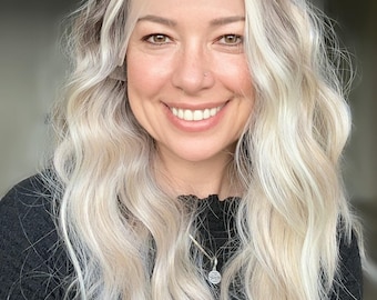 May include: A woman with long, wavy, platinum blonde hair is wearing a black, short-sleeved shirt. She is smiling and has a silver necklace with a small, round pendant.