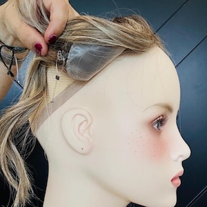 May include: A mannequin head displaying a wig, viewed in profile. The wig combines blonde and light brown strands. The mannequin has painted facial features, including freckles and blush. The wig's lace front is visible.