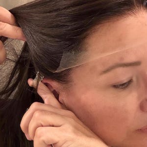 May include: A close-up of a person's head and shoulder, with dark brown hair. A lace front wig is being applied, with the lace visible at the hairline. The person's hand is holding the wig in place.