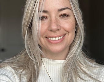 May include: A smiling person with blonde hair and a white ribbed sweater. A silver necklace with a large, dark blue and brown pendant hangs around their neck. The person is looking directly at the camera.