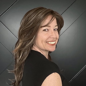 May include: A woman with long, layered brown hair with blonde highlights smiles at the camera. She is wearing a black top. The background is a dark gray wall with diagonal lines.