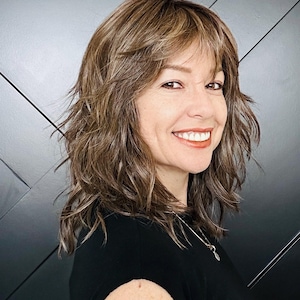 May include: A shoulder-length brown wig with layered texture and wispy bangs. The hair features multi-tonal highlights in shades of light brown and caramel, styled with a soft, wavy finish that frames the face and adds volume to the overall appearance.