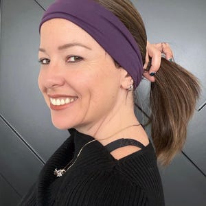 May include: A woman wearing a dark purple headband and a black sweater. The headband is wide and covers the forehead. The woman is smiling and has brown hair pulled back in a ponytail. She is wearing a silver necklace.