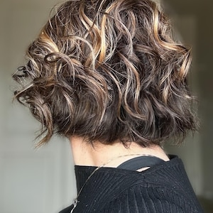 May include: A close-up of a person's short, wavy hairstyle. The hair is a mix of dark brown, light brown, and blonde highlights. The hair is styled in a layered, textured bob. The person is wearing a black top and a silver necklace.