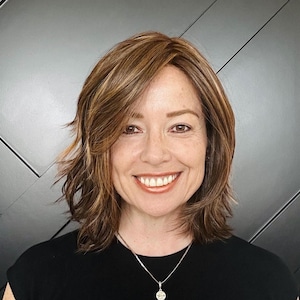 May include: A woman with a warm smile, wearing a black top and a silver necklace with a pendant. She has layered, shoulder-length brown hair with highlights. The background is a textured gray wall.