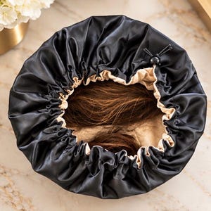 May include: A circular hair bonnet featuring a smooth black outer layer and a beige inner lining. The bonnet has a gathered, elasticized edge with a black adjustable drawstring toggle closure, containing a section of brown hair inside the center.