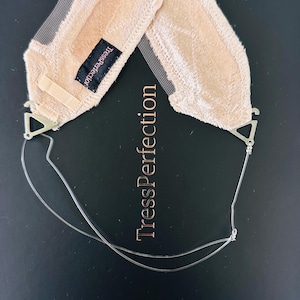 Puede incluir: Una banda de sujeción para peluca de encaje beige con un centro de malla y correas ajustables. El producto tiene clips metálicos y una pequeña etiqueta negra que dice "TressPerfection". El texto "TressPerfection" también está impreso en el fondo negro.