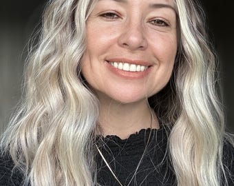 May include: A smiling person with long, wavy blonde hair and a black top. A silver necklace with a small, round pendant hangs around their neck. The background is dark.