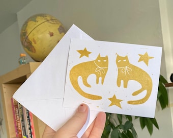 Op de afbeelding: Een wenskaart met twee gestileerde gouden katten, strikjes en sterren op een witte achtergrond. De kaart wordt voor een witte envelop gehouden. Een wereldbol en boeken zijn zichtbaar op de achtergrond.