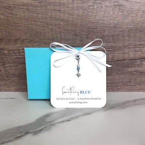 May include: A white card with the text "Something BLUE" and a charm with blue beads and a star. The card is tied with a white ribbon and sits on a blue box. The text below the title reads "for love so true - a timeless thread in something new."