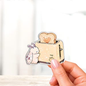 May include: A die-cut sticker featuring a light yellow toaster with a slice of bread popping out, which has a heart-shaped center. A small, fluffy white moth with pink-spotted wings stands beside the toaster. The word toast is printed on the front.