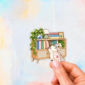 May include: A die-cut sticker featuring a light-colored moth with pink-spotted wings standing next to a wooden cabinet. The cabinet holds books, a lamp, a potted plant, a snow globe, and a framed picture of a lily of the valley flower.